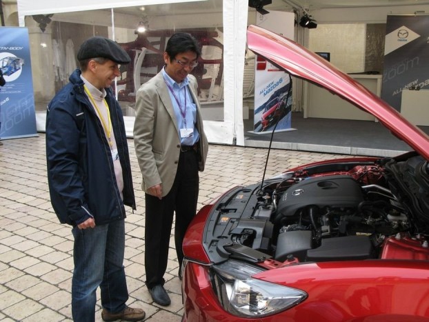 Vezettük az új Mazda6-ost! 5 Kajiyama szannal, az új Mazda6 atyjával, vagy ahogy gyári berkekben nevezik, programmenedzserével nézegetjük a motorteret. Utána megmutatta a gyalogosbarát, összeomló motorháztető zsanért, amire nagyon büszke. Igénytelen megoldás a pálcás kitámasztás